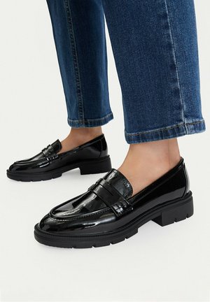 Person wearing shiny black leather loafers and blue jeans standing on a light surface, focusing on footwear and lower legs.