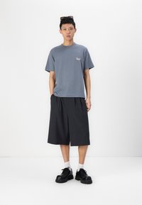 Sivá bavlnená tričko s malým logom, v kombinácii s tmavými, krátkymi nohavicami po kolená. Čierne masívne topánky a biele ponožky dotvárajú outfit.
