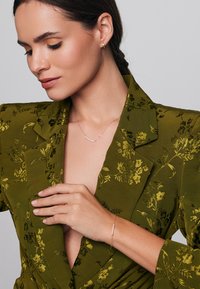 Zielona marynarka w kwiatowy wzór z fakturowanej tkaniny. Modelka nosi delikatny złoty naszyjnik i bransoletkę z małymi kamieniami szlachetnymi, które podkreślają minimalistyczny styl.