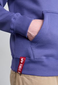 Violetinis džemperis su banguotomis rankovėmis ir apatine krašto juosta, turintis kišenę ir raudoną etiketę su užrašu "PAŠALINTI", esantį apatinio siūlės šone.