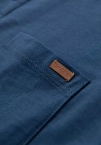 Tela de algodón azul marino con un bolsillo de parche. Presenta una etiqueta de cuero marrón con el texto "ombre", con un detalle de costura visible.
