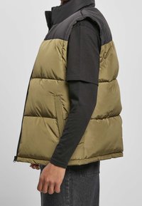 Pufferväst med en svart övre del och en olivgrön nedre del. Har dragkedjestängning, sidofickor och en lätt, vadderad struktur.