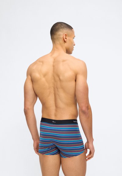 Άνδρας με κοντά μαλλιά που δείχνει μυώδη πλάτη, φορώντας μπλε μποξεράκια με πολύχρωμες ρίγες και μαύρη μέση.