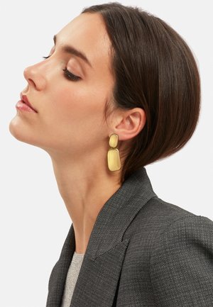 Gouden oorbellen met een gladde, rechthoekige vorm en een gepolijste afwerking. Draagt door een persoon in een grijze blazer, die hun silhouet benadrukt.