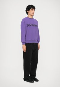 Lilla sweatshirt med teksten "No Problem" i sort. Har ribbede manchetter og kant, parret med sorte baggy bukser og sorte sneakers.