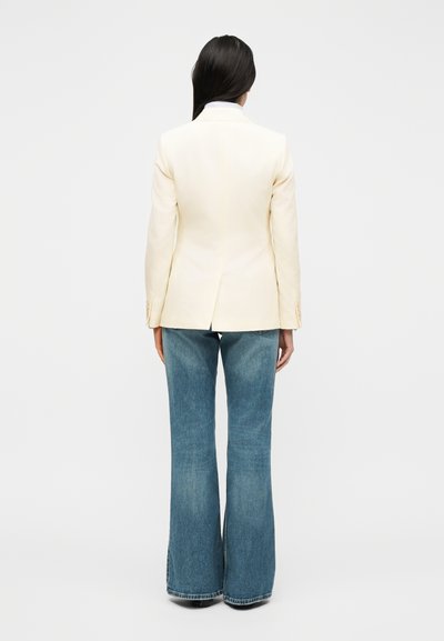 Γυναίκα με μακριά σκούρα μαλλιά που φοράει εκρού σακάκι και καμπανιστά μπλε τζιν, στέκεται με την πλάτη προς το μέρος μας σε ένα απλό φωτεινό φόντο.