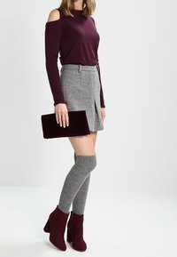 Burgunderfarbendes Top mit kalten Schultern, grauer karierten Minirock, kniehohe graue Socken und burgunderfarbene Stiefeletten, die eine burgunderfarbene Samt-Clutch hält.