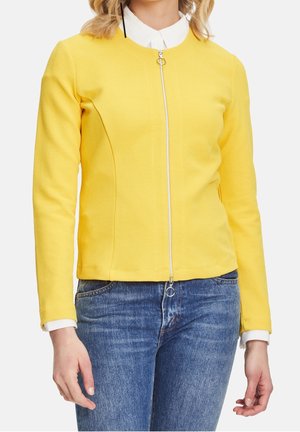 Kobieta ubrana w dopasowaną żółtą kurtkę na zamek błyskawiczny na białą koszulę z kołnierzykiem, w połączeniu z niebieskimi jeansami, pokazana od szyi do bioder.