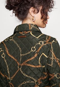 Quiltad grön jacka med guldchain och brunt remmönster. Har en krage och en metalldetail på den övre delen av ryggen.