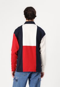 Camisa polo de manga larga con un diseño a bloques de color en azul marino, rojo y crema. La tela es de algodón con un acabado texturizado. Cuello clásico y mangas con puños.