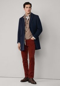 Marineblauer Mantel über einem beigen Cardigan und karierten Hemd, kombiniert mit rostfarbenen Hosen und braunen Lederstiefeln, vor einem neutralen Hintergrund.