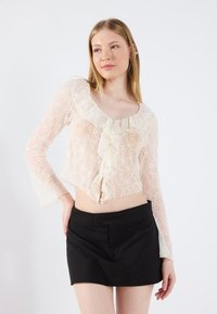 Blonde topp i lys krem med bølget halsringning og klokkeermer, parret med en svart miniskjørt med glatt tekstur og tettsittende design.