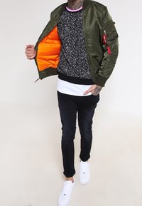 Veste bomber vert olive avec doublure orange, pull noir moucheté, t-shirt blanc, pantalon foncé et baskets blanches. Détail avec poche sur la manche.