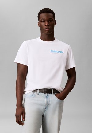 Młody mężczyzna w białej koszulce Calvin Klein z niebieskim logo, jasnoniebieskich dżinsach i czarnym pasku, stojący na jednolitym szarym tle.