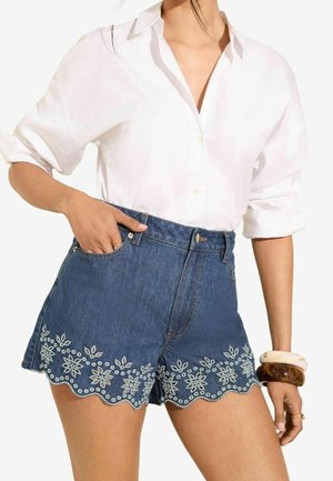 Femme portant une chemise blanche boutonnée avec les manches retroussées et un short en jean taille haute avec un ourlet festonné et une broderie florale blanche.