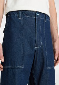 Pantalones de mezclilla en un tono azul oscuro con corte recto. Presentan bolsillos frontales y traseros, costuras en contraste y cierre de botón.