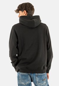 Felpa nera con cappuccio ampio, polsini a costine e orlo a costine. Indossata sopra jeans di denim azzurro chiaro, con tasche posteriori caratterizzate da dettagli di cucitura.