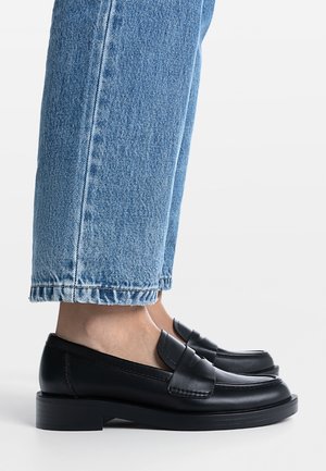 Pieds chaussés de mocassins en cuir noir associés à un jean denim bleu raccourci au-dessus des chevilles sur un fond clair uni.