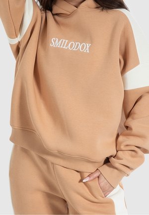 Beiges Hoodie- und Sweatpants-Set mit weißen Akzenten, mit dem Text "SMILODOX" auf der Brust und einer Hand in der Tasche.