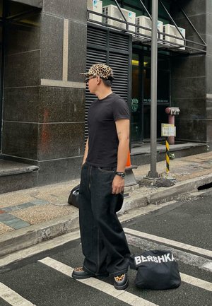 Uomo con t-shirt nera e jeans larghi, che indossa un cappellino e scarpe con stampa leopardata, in piedi per strada accanto a una borsa nera con la scritta "BERSHKA".