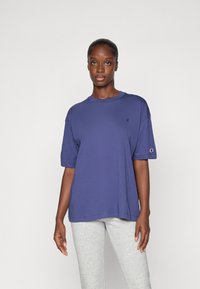 T-shirt in cotone viola con collo tondo, maniche corte e un piccolo logo sul lato sinistro del petto. Abbinato a pantaloni della tuta grigio chiaro.