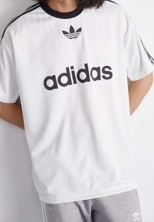 Osoba nosząca białą koszulkę Adidas z czarnym logo i paskami, w połączeniu z szarymi szortami Adidas z białym logo.