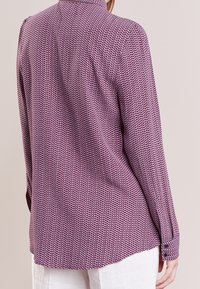 Camicetta a maniche lunghe con un motivo a zigzag viola e bianco, con polsini con bottoni e un orlo leggermente curvato. Tessuto leggero.
