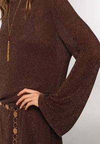 Top de punto metálico marrón con mangas largas acampanadas, que presenta un acento de cinturón de círculo dorado y un colgante largo de borlas doradas. Texturizado con un elegante brillo.