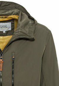 Veste vert olive avec une fermeture éclair à l'avant, doublure en maille jaune et deux poches zippées. Comprend une capuche et des détails matériels contrastés.