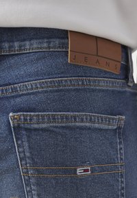 Gros plan de la poche arrière d'un jean en denim bleu avec un patch en cuir marron marqué « JEANS » et un petit logo drapeau sur la couture de la poche.