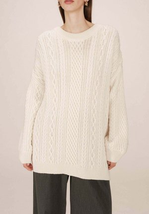 Pull en maille torsadée crème avec un col rond, des motifs texturés contrastants et de longues manches généreuses, associé à un pantalon large gris.