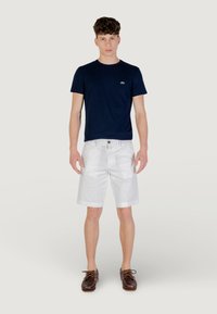 T-shirt en coton bleu marine à manches courtes avec un petit logo, associé à un short blanc et des chaussures bateau marron, se tenant devant un fond neutre.