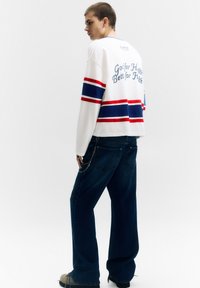 Fehér pulóver kék és piros csíkokkal, valamint felirattal a háton. Sötétkék bőszárú farmernadrághoz és bézs cipőhöz párosítva. Egyszínű háttér.