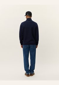 Navy pullover with high collar, ribbed cuffs, and hem. Worn with blue denim jeans and brown shoes, viewed from the back.