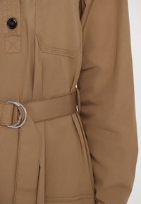 Cappotto beige con cintura, dotato di tasca con bottone, texture liscia e vita arricciata con fibbia in tonalità argento.