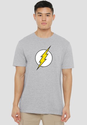 Junger Mann trägt ein hellgraues T-Shirt mit einem gelben Blitz in einem weißen Kreis auf der Brust und steht vor einem einfarbigen Hintergrund.