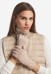 Guanti in maglia di un morbido beige con polsini a coste. Il modello indossa un gilet imbottito beige e un dolcevita bianco, con le mani delicatamente unite.
