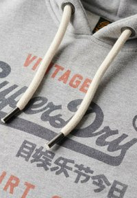Grå hoodie har en snor med hvide strukturerede kord og metalliske ender, der viser et vintagemotiv i forskellige farver på forsiden.