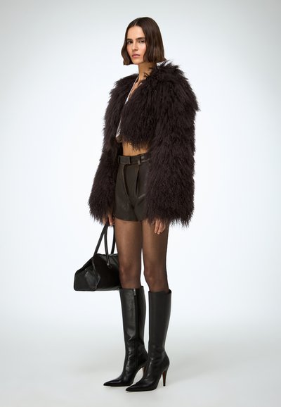 Zwarte nep bont cropped jas over een beige top, gecombineerd met bruine leren shorts, zwarte kniehoge laarzen met hak en een zwarte handtas.