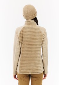 Beige fleecjas met een gestructureerde achterkant, gladde mouwen en een hoge kraag. Gecombineerd met een bijpassende beige gebreide muts.