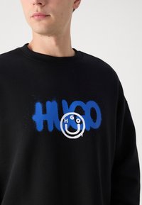 Crna dukserica s plavim grafitnim tekstom koji glasi "HUGO" i kružnim logotipom s "HGO" i dizajnom s osmijehom.