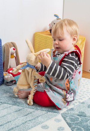 Bambino seduto su un tappeto decorato, che tiene e osserva un coniglio di peluche beige in una stanza giochi con altri peluche nelle vicinanze.