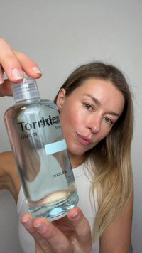 Woman with long hair holding a clear bottle labeled "Torriden Dive In Toner" containing low molecular hyaluronic acid, against a plain background.