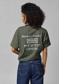 Olīvzaļš lielizmēra T-krekls ar baltu drukātu tekstu: "RED BULL RACING," "RBR," un "PIT STOP CREW." Saskaņots ar tumši ziliem džinsiem.
