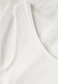 Top sin mangas de algodón blanco con textura suave, escote amplio y detalles de costura limpios, colocado sobre una tela similar.