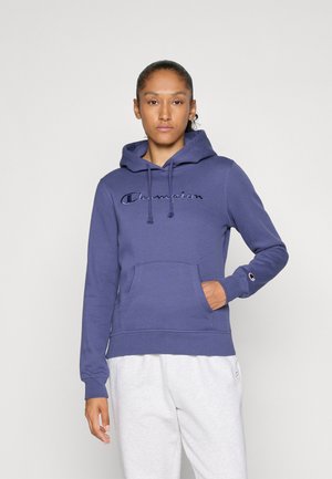 Kvinne iført en blå Champion hettegenser med logo på brystet og ermet, kombinert med hvite joggebukser, som står mot en ensfarget bakgrunn.