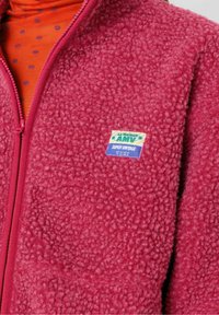 Flis jakna v temno roza barvi z teksturirano površino; ima zadrgo in barvno znamčno oznako na prsih.