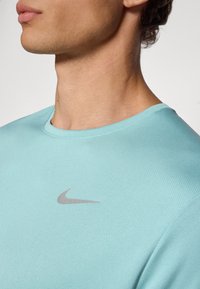 Világoskék sportpóló texturált anyagból. Kerek nyakkivágással és egy kis szürke Nike logóval rendelkezik a mellkason.