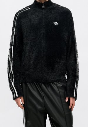Person iført en sort, blød lynlåstrøje med hvide Adidas-striber på ærmerne og logo på brystet, kombineret med sorte læderbukser.