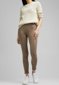Vrouw in een beige gebreide trui, bruine strakke broek en witte sneakers, staand tegen een effen grijze achtergrond.